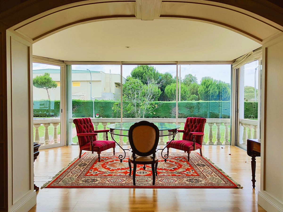 A bright and airy sitting area features expansive glass windows that provide views of the surrounding greenery. A round table is surrounded by elegant chairs, while a patterned area rug adds warmth to the wooden floor.