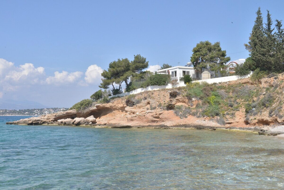 A traditional house is situated on a peninsula, surrounded by clear waters. The property features a coastal landscape with rocky shores and a backdrop of green trees, enhancing the natural scenery. Soft waves gently lap at the shoreline.