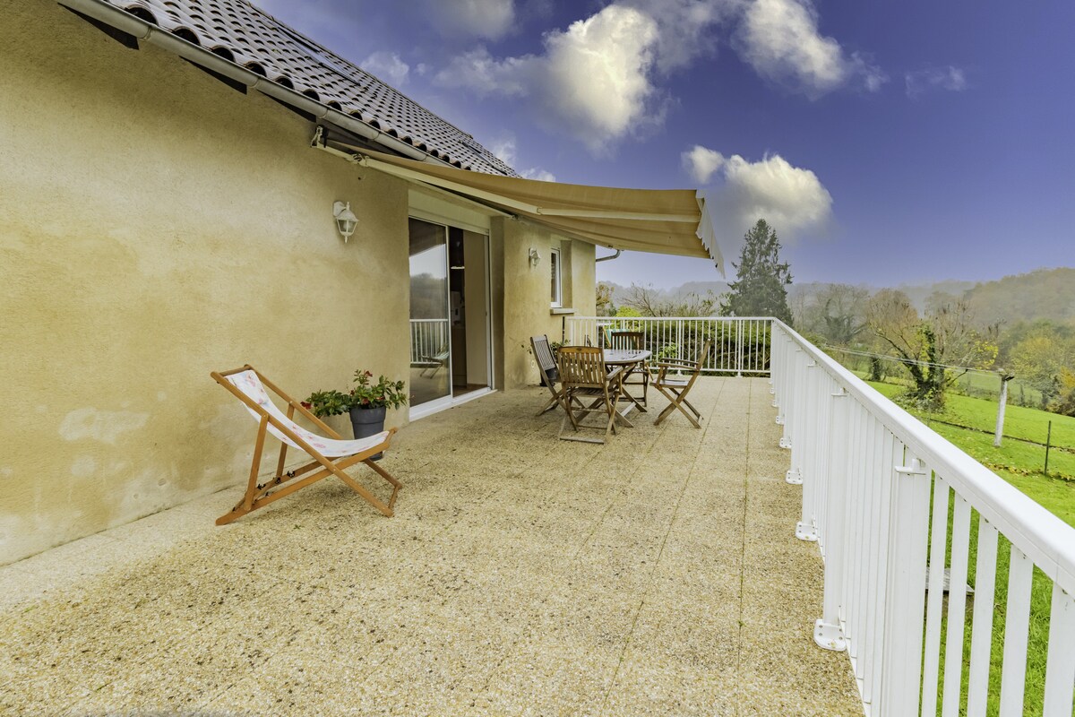 A spacious terrace features a light-colored stone surface with a wooden foldable chair positioned for relaxation. A small table with two chairs is visible, and a potted plant adds a touch of greenery. The surrounding view includes grasslands and distant trees under a cloudy sky.
