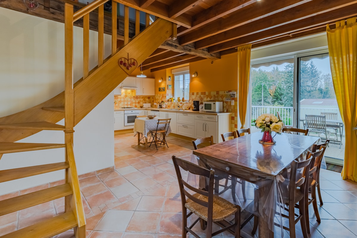 A spacious living area is featured, showcasing a wooden dining table with chairs, surrounded by soft yellow curtains. A modern kitchen is visible, equipped with essential appliances. A staircase leads to the upper level, complementing the inviting atmosphere and open layout.