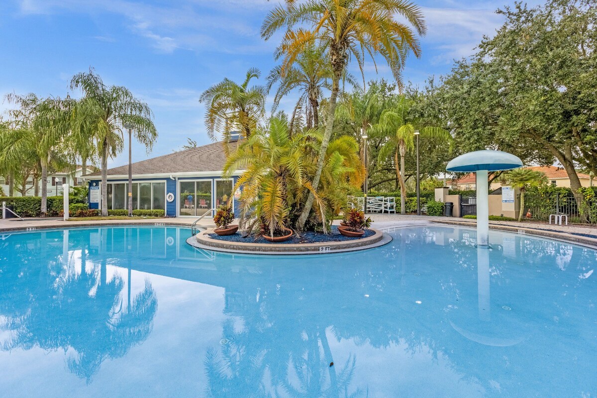 A large, inviting pool area is surrounded by tropical palm trees and colorful landscaping. A shaded seating area is visible nearby, and a cabana offers additional shaded space. The water reflects the clear blue sky, creating a serene atmosphere for relaxation.