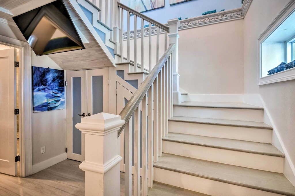 A well-lit staircase features white wooden banisters and light-colored steps leading to an upper level. A decorative painting hangs on the wall near the bottom of the stairs. Natural light filters in through a window at the landing, enhancing the open feel of the space.