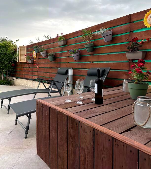 A back garden area features a wooden table with an elegant arrangement of wine glasses and a bottle. Two lounge chairs are positioned nearby, complemented by a wooden wall adorned with various potted plants, creating a serene and inviting outdoor space.