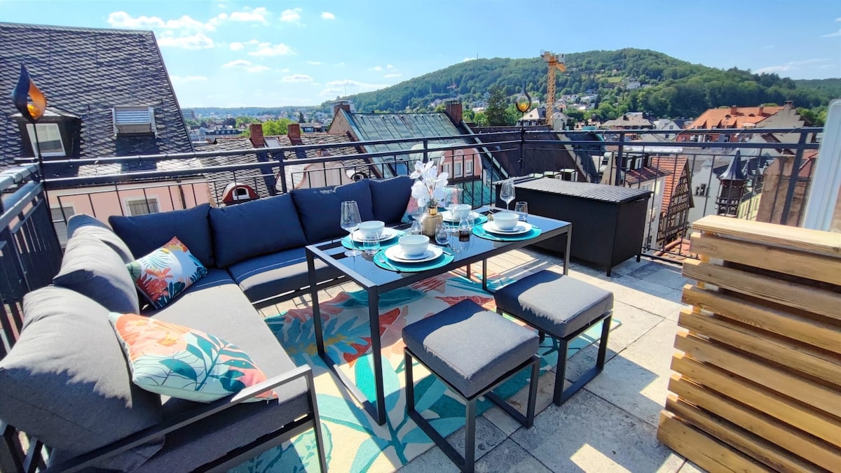 A spacious rooftop terrace is shown, furnished with an inviting seating arrangement, including a large table surrounded by cushioned chairs. Decorative table settings are arranged atop the table, and a vibrant area rug adds color. Scenic views of the surrounding hills are visible in the background.