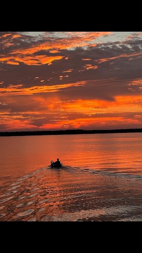 A sunset scene is captured over a tranquil lake, with hues of orange and pink reflecting on the water's surface. A small boat is seen gently moving across the lake, creating ripples in the water. The sky features scattered clouds illuminated by the setting sun.