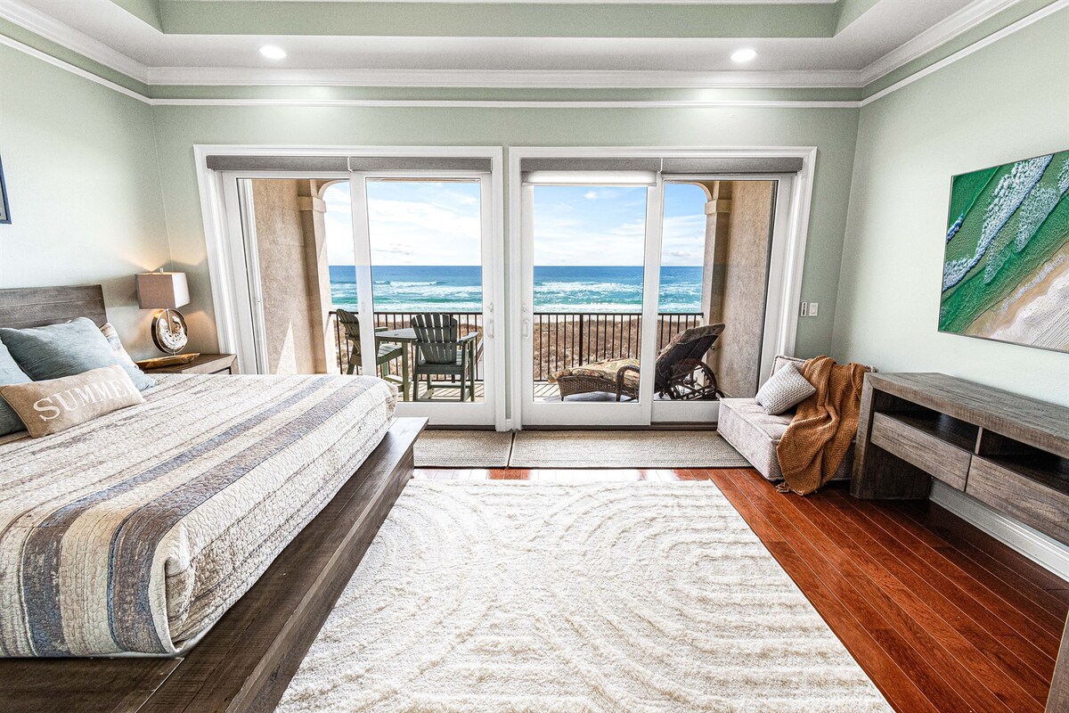 A spacious bedroom features large sliding glass doors that open to a private balcony overlooking the beach. A comfortable bed lies against one wall, complemented by a cozy seating area with a chair. A neutral-toned area rug covers the hardwood floor, adding warmth to the space.