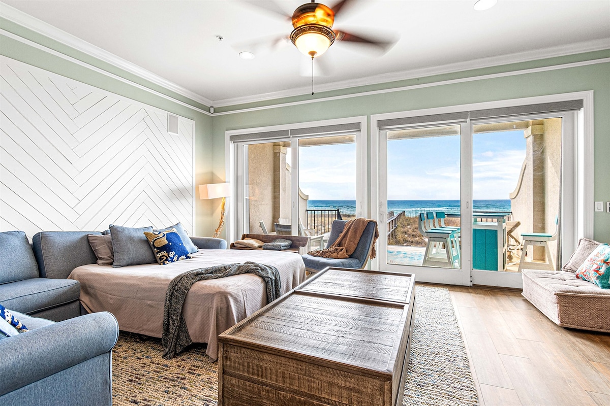 A light-filled living area features a large sectional sofa and a spacious bed, complemented by a textured rug. Floor-to-ceiling sliding doors open to a private patio with ocean views. Natural light enhances the calming color scheme, creating a welcoming space for relaxation.