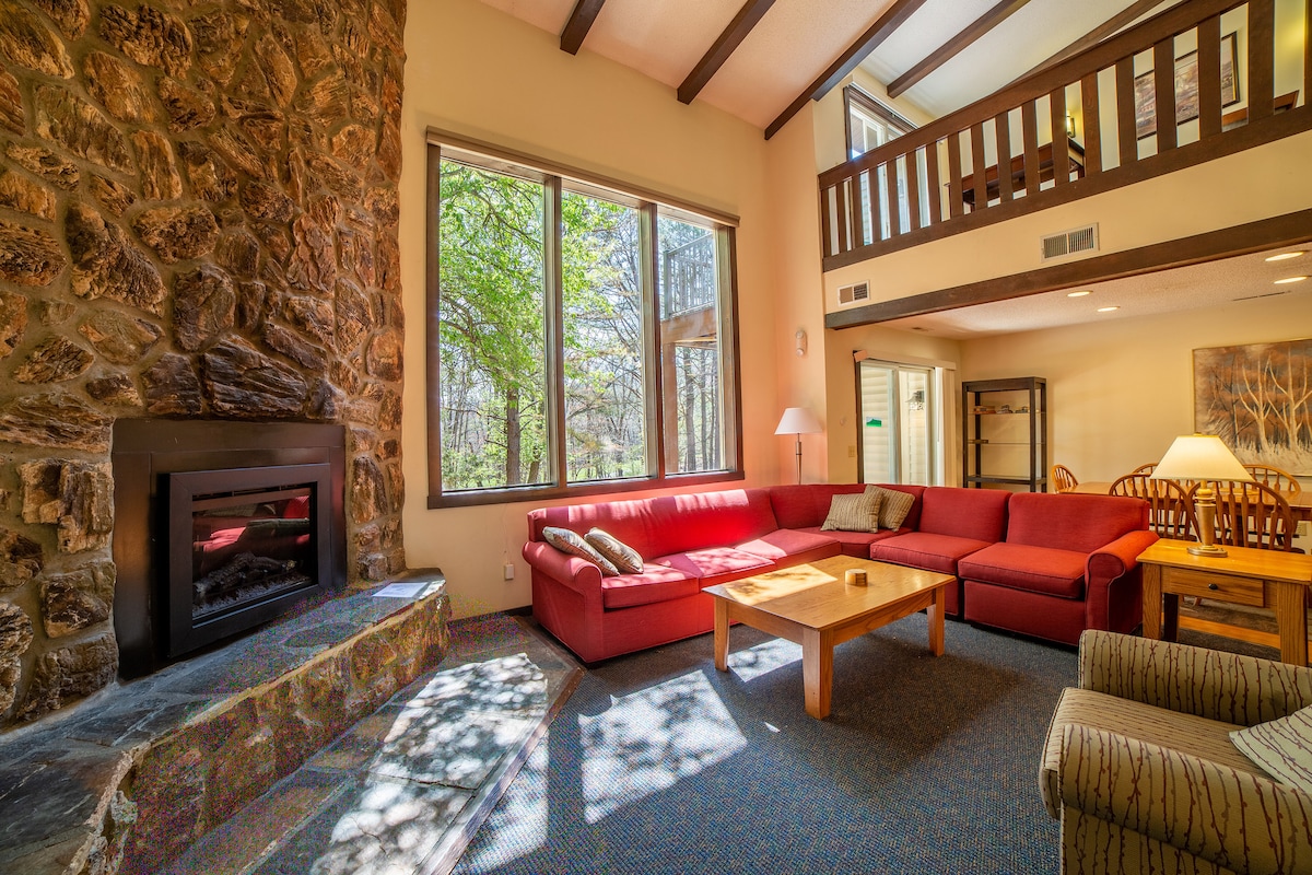A spacious living room features a large red sectional sofa and a wooden coffee table. A stone fireplace is situated against one wall, while expansive windows provide natural light and views of the surrounding trees. A loft area above adds an open feel to the space.