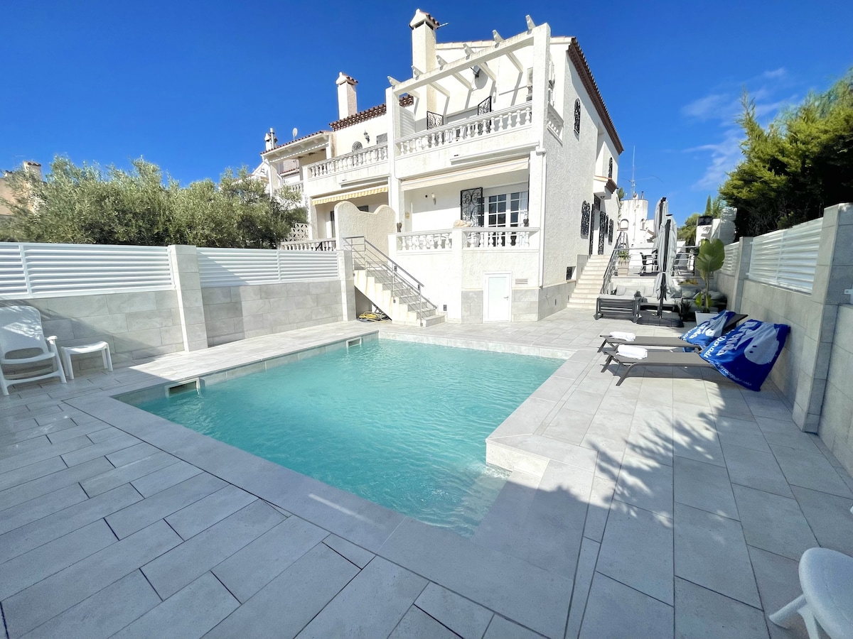 A private pool is surrounded by a spacious patio area, featuring light-colored paving and a mix of sun loungers and chairs. The villa's two-story structure is visible in the background, along with greenery and outdoor steps leading to the upper level.
