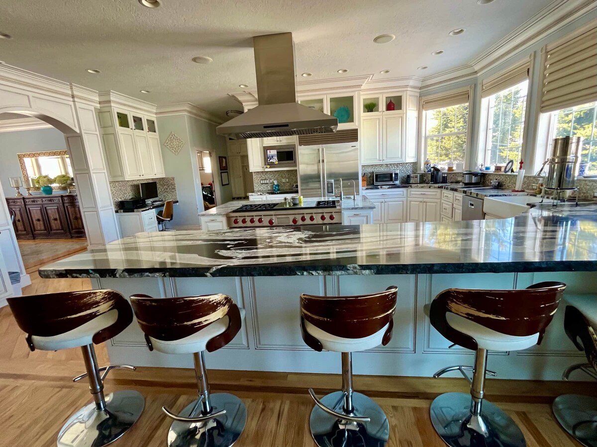 An industrial kitchen displays a spacious marble countertop with seating for several guests at sleek bar stools. Bright natural light fills the area through large windows, highlighting the modern appliances and cabinetry in a classic white finish.