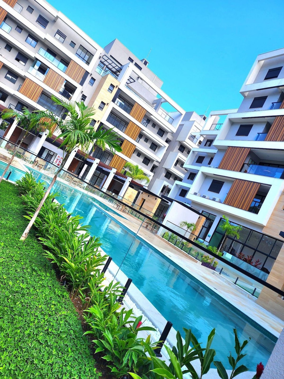 A modern condominium complex is shown, featuring multiple buildings set around a tranquil pool area. Lush greenery lines the pathways, and clear blue waters reflect the bright sky. Spacious outdoor living spaces blend seamlessly with the architecture, creating an inviting atmosphere for relaxation.