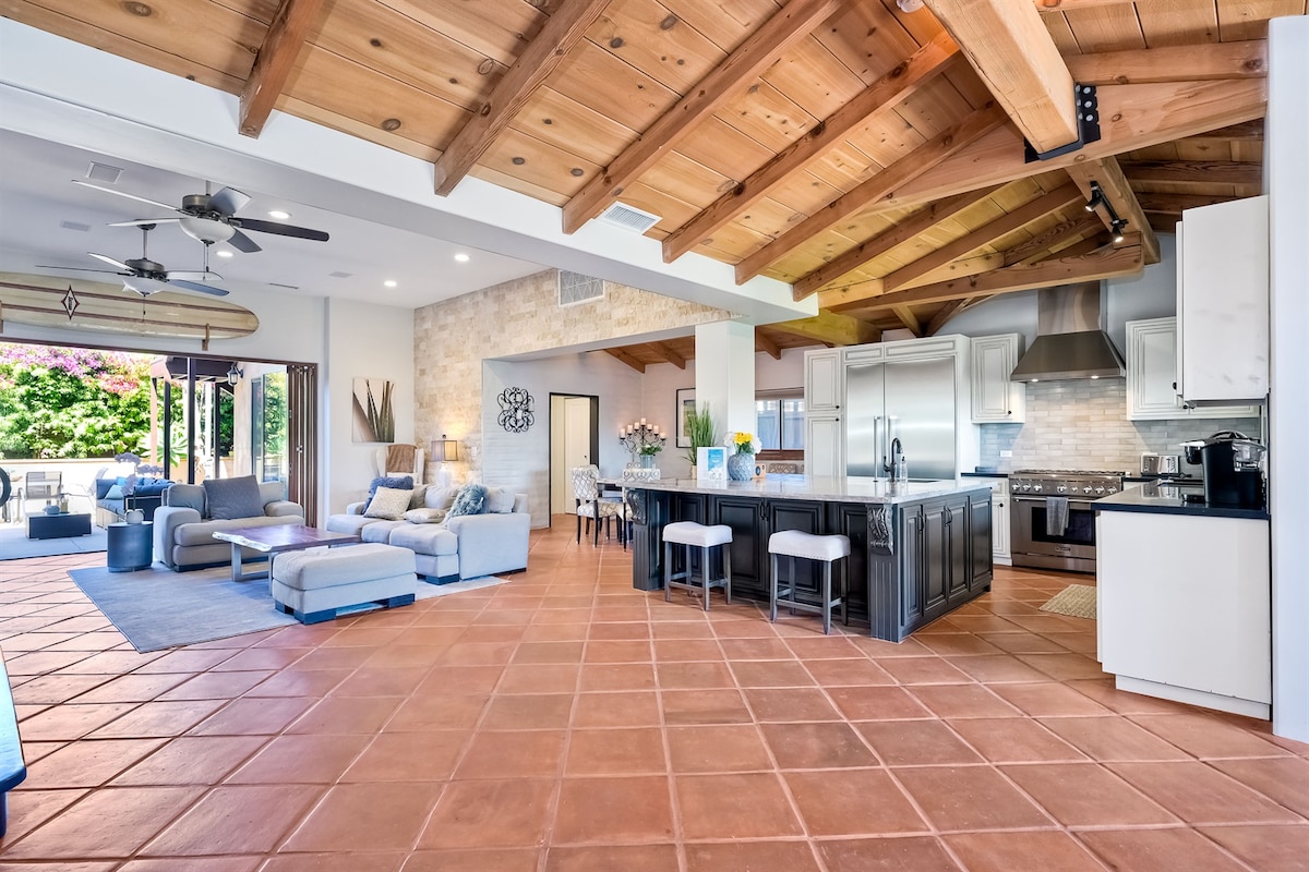A spacious living area features a high ceiling with wooden beams and terracotta tile flooring. The area includes comfortable seating with light-colored furniture and a smart TV. A modern kitchen with an island and sleek appliances is visible, enhancing the open layout.