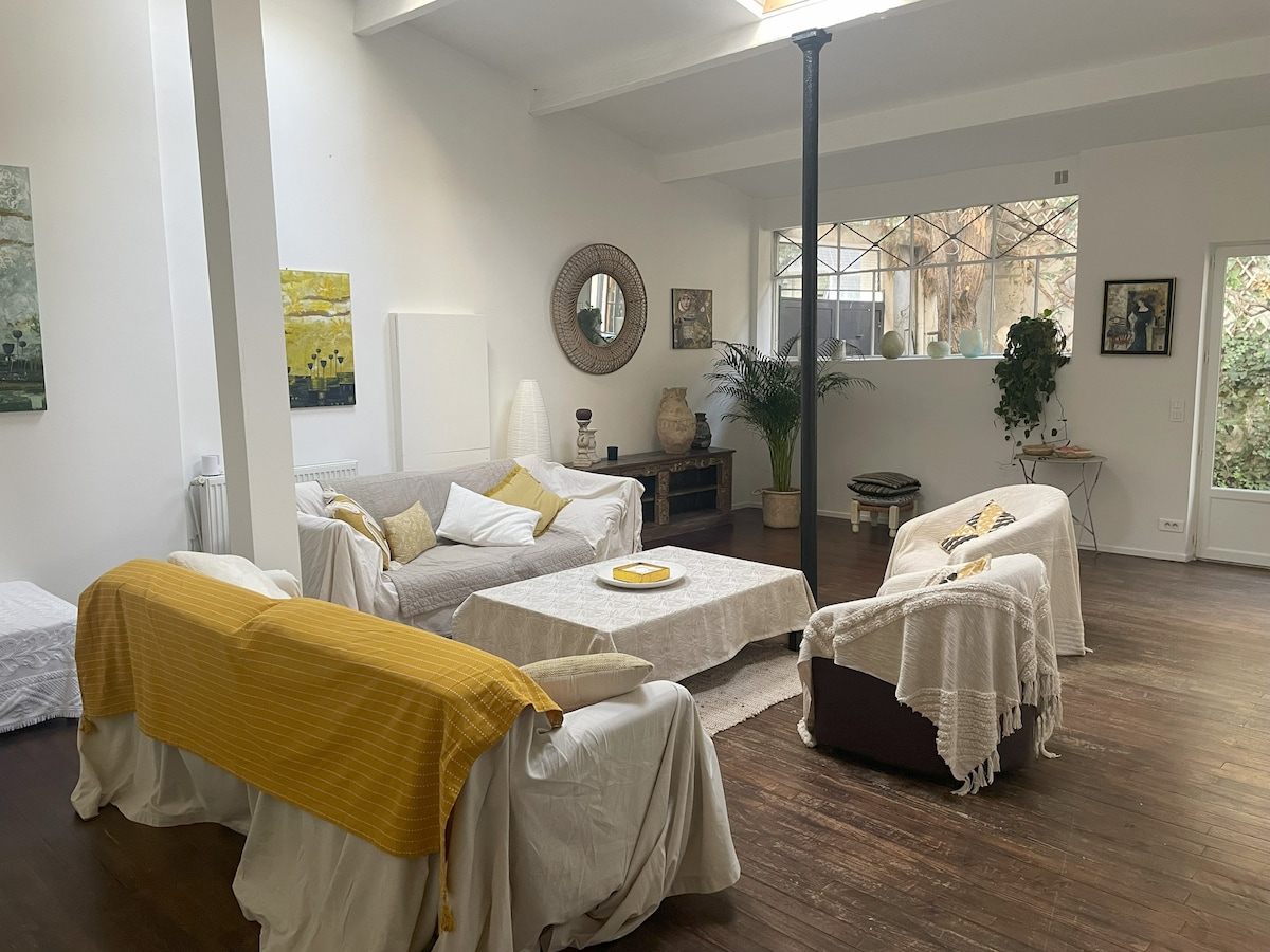 A spacious living area features comfortable seating arrangements with light-colored sofas draped in soft throws. Natural light pours in through large windows, illuminating the wooden floor and various decorative elements, including potted plants and art pieces that enhance the serene atmosphere.