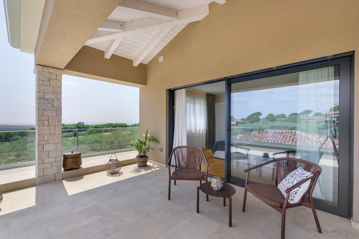 A covered patio is featured, showcasing two wicker chairs with cushions and a small table. Floor-to-ceiling glass doors provide a view into the interior space while framing scenic outdoor greenery. A potted plant adds a touch of nature to the inviting area.