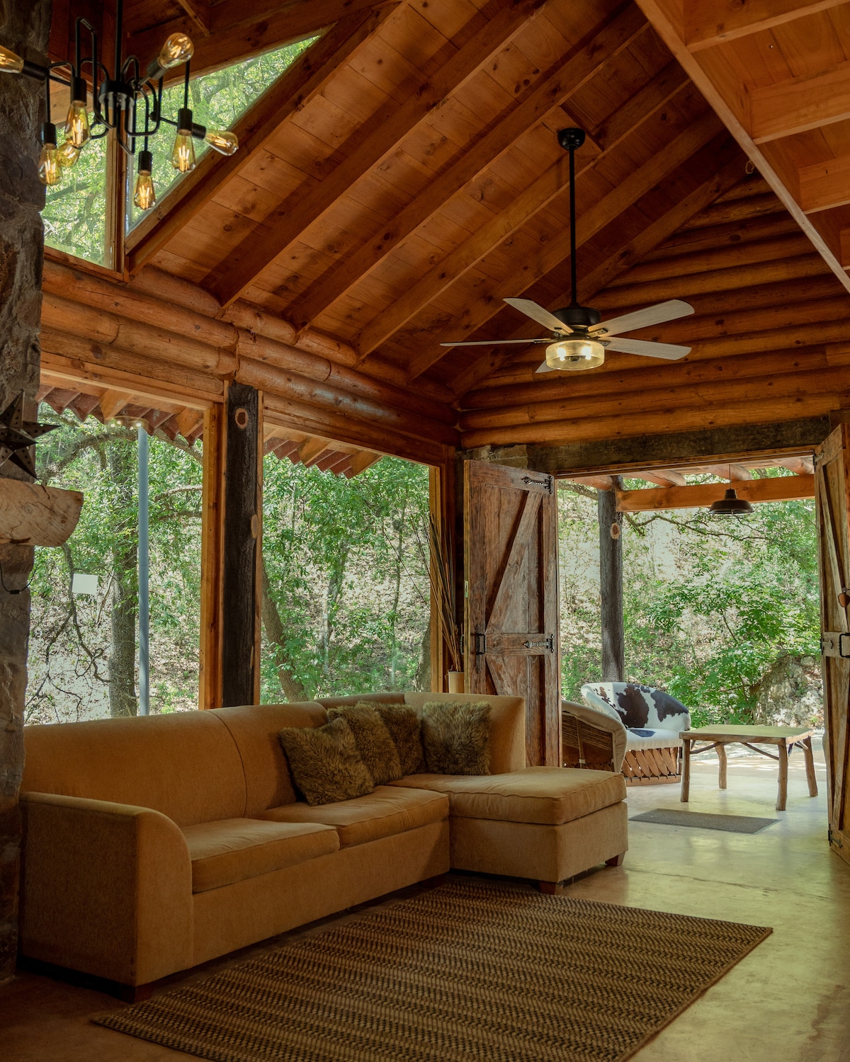 A welcoming living area features a cozy sectional sofa adorned with accent pillows. Large windows provide a view of the surrounding greenery, allowing natural light to fill the space. The wooden ceiling adds warmth, while a ceiling fan enhances airflow.