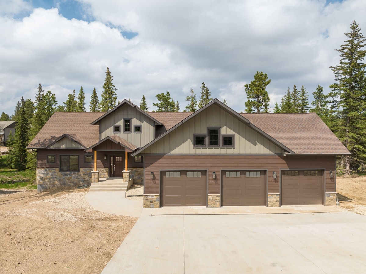 A newly completed cabin is presented, featuring a combination of stone and wood siding. The property includes a three-car garage with large doors. Surrounding the home are lush evergreen trees under a partly cloudy sky, providing a natural backdrop.