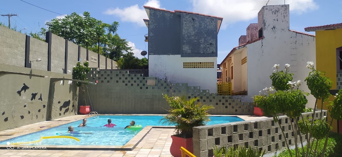 A communal swimming pool area is surrounded by tall walls covered in artful designs. Guests can be seen enjoying the water, while lush greenery and decorative plants enhance the space. Bright sunlight reflects off the pool's tranquil blue water, inviting relaxation.