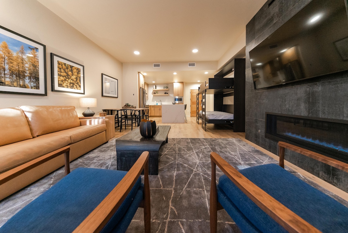 The living area features a combination of a leather sofa, blue upholstered chairs, and a coffee table. A wall-mounted TV is situated above a modern fireplace. The space is illuminated by natural light, with a view of the dining area and kitchen in the background.