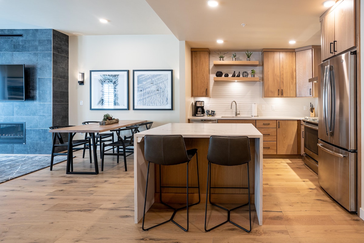 A modern kitchen and dining area are visible, featuring a spacious island with two bar stools. Stainless steel appliances and wood cabinetry highlight the kitchen. A dining table with six chairs is set beside the kitchen, complemented by artwork on the wall.