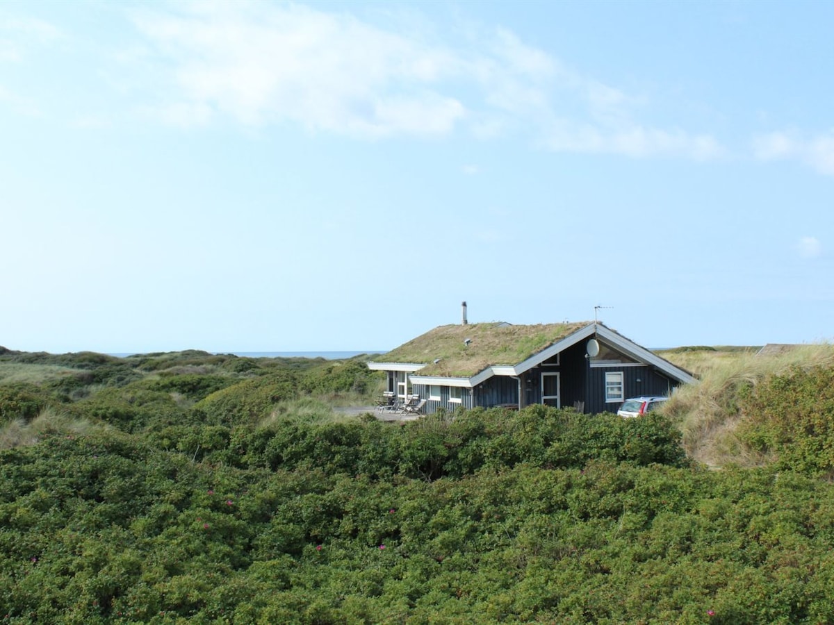 A single-family house with a green roof is surrounded by lush greenery, blending into the natural landscape. The ocean is visible in the distance, and a terrace area is included beside the structure. The building features a traditional design with large windows.