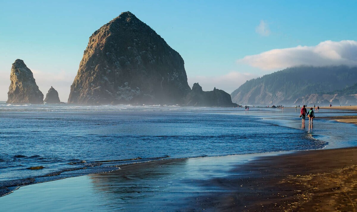 A tranquil beach scene features gentle waves lapping at the shore, with rocky formations rising dramatically from the water. Several figures can be seen walking along the wet sand, with soft blue skies and distant hills providing a serene backdrop.