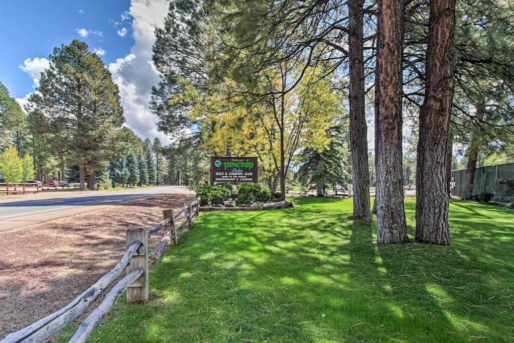 A welcoming sign for Pinetop is displayed amidst lush green grass and towering pine trees. The surroundings reflect a serene outdoor setting, with a clear blue sky overhead, suggesting a peaceful atmosphere for visitors. Wooden fencing lines the area, enhancing the natural beauty.