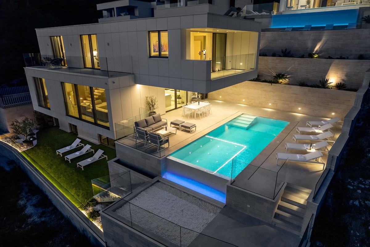 An aerial view showcases the villa's modern architecture, highlighted by a spacious, illuminated infinity pool surrounded by deck chairs. A cozy outdoor lounge area and dining table are visible, enhanced by soft outdoor lighting, amidst tiered landscaping that complements the property.
