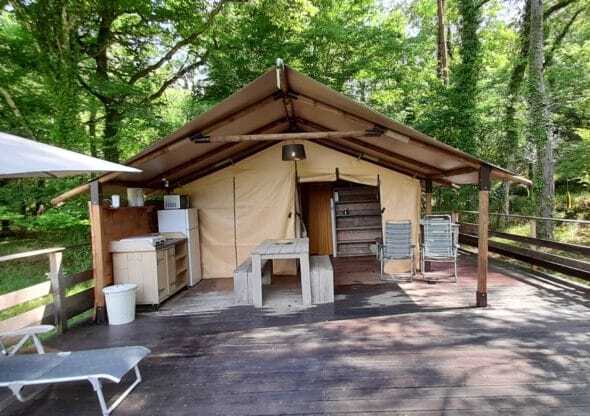 A spacious wooden deck extends from the tent, featuring outdoor dining furniture and two lounge chairs. The equipped kitchen area is visible, with a refrigerator and countertops under a shaded awning, surrounded by lush green trees enhancing the natural setting.