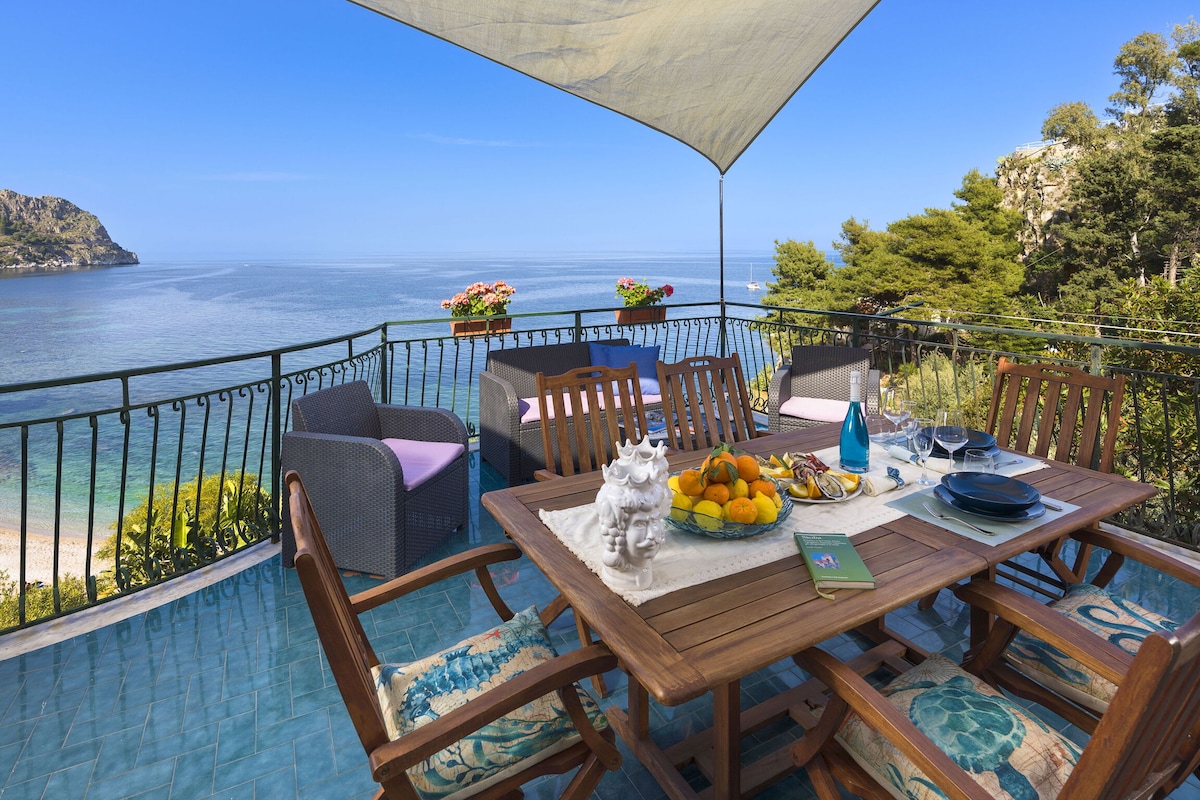 A spacious terrace boasts a dining table surrounded by wooden chairs. Sunlight reflects off the blue water, while potted flowers add color. A shaded area offers comfort with a view of the sea and nearby cliffs, creating an ideal setting for outdoor meals.