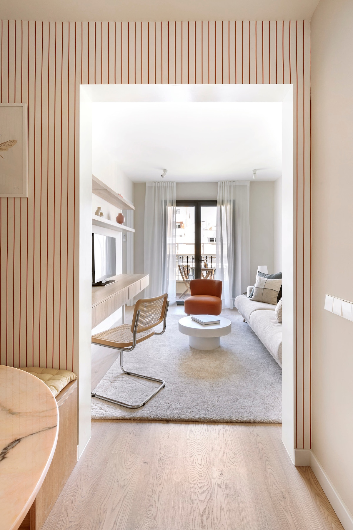 An inviting hallway leads into a well-lit living area furnished with a cozy sofa and a comfortable accent chair. Neutral tones are complemented by soft textiles, while large windows allow natural light to illuminate the space. Subtle decor elements enhance the modern design.