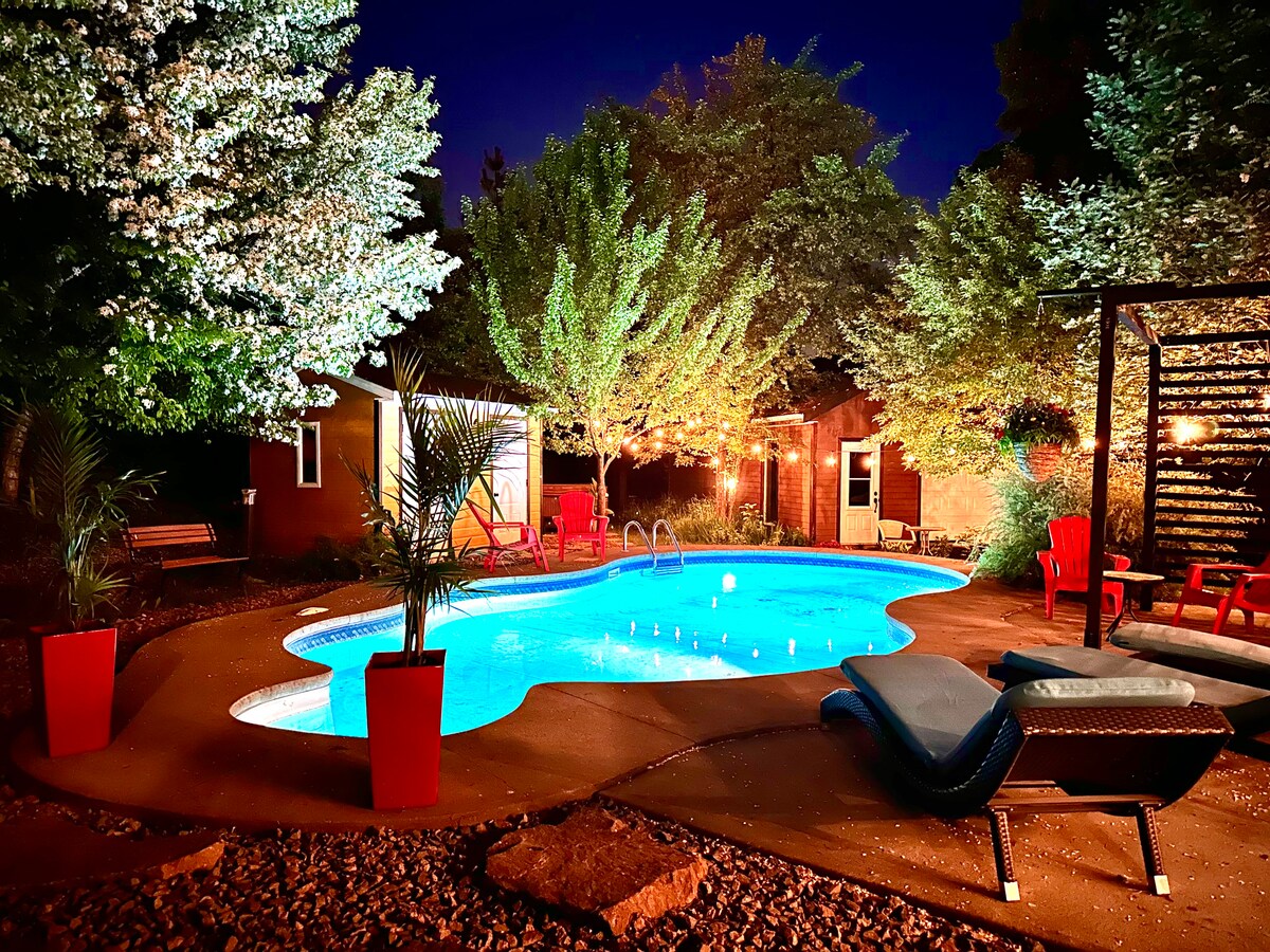 An inviting outdoor space is illuminated by soft lighting, featuring a swimming pool surrounded by lush greenery. Red chairs and planters add vibrant color, while a lounge chair beside the pool offers a relaxing spot. The ambiance is warm and serene, perfect for unwinding.