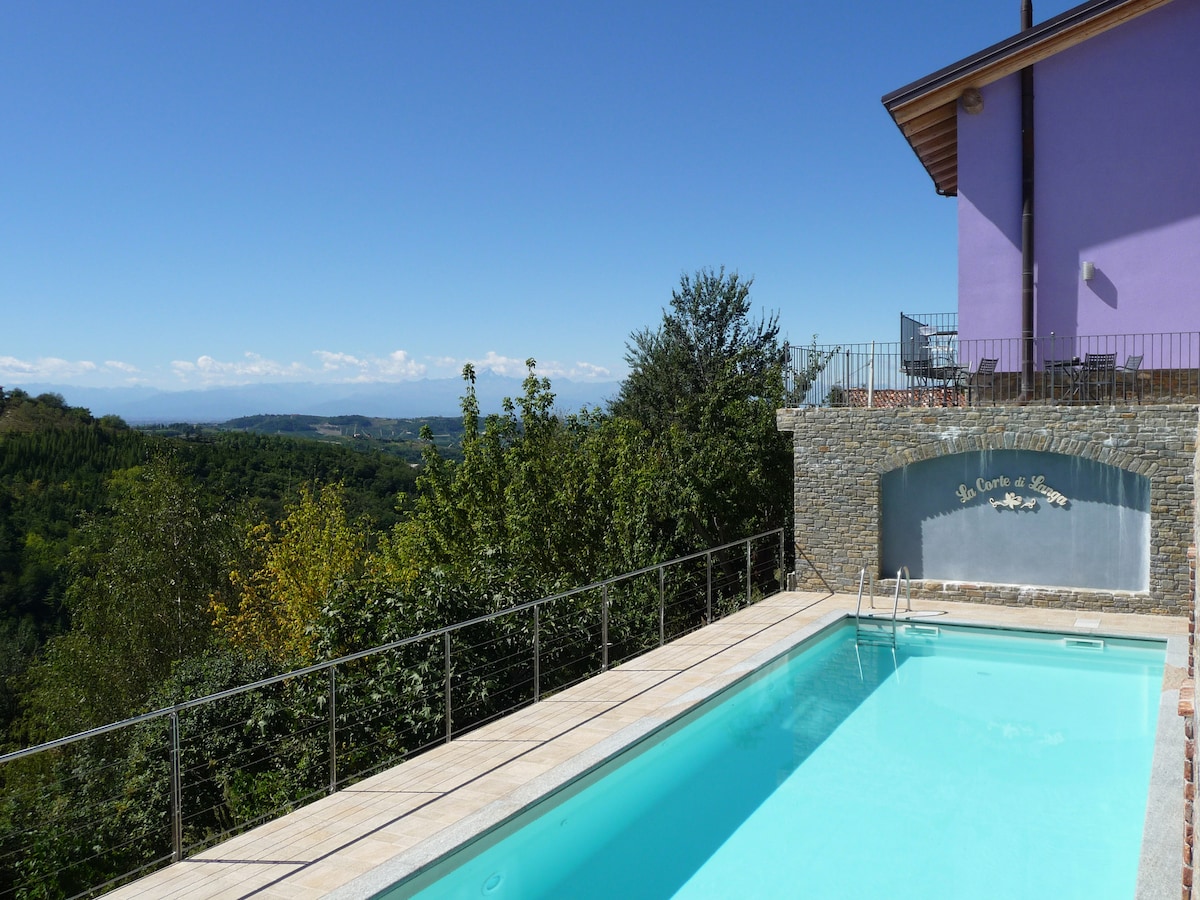A serene view of a shared outdoor swimming pool is presented, bordered by a smooth stone deck. Lush greenery surrounds the area, with distant hills visible under a clear blue sky. The property is partially visible in the background, featuring a gently sloping roof.