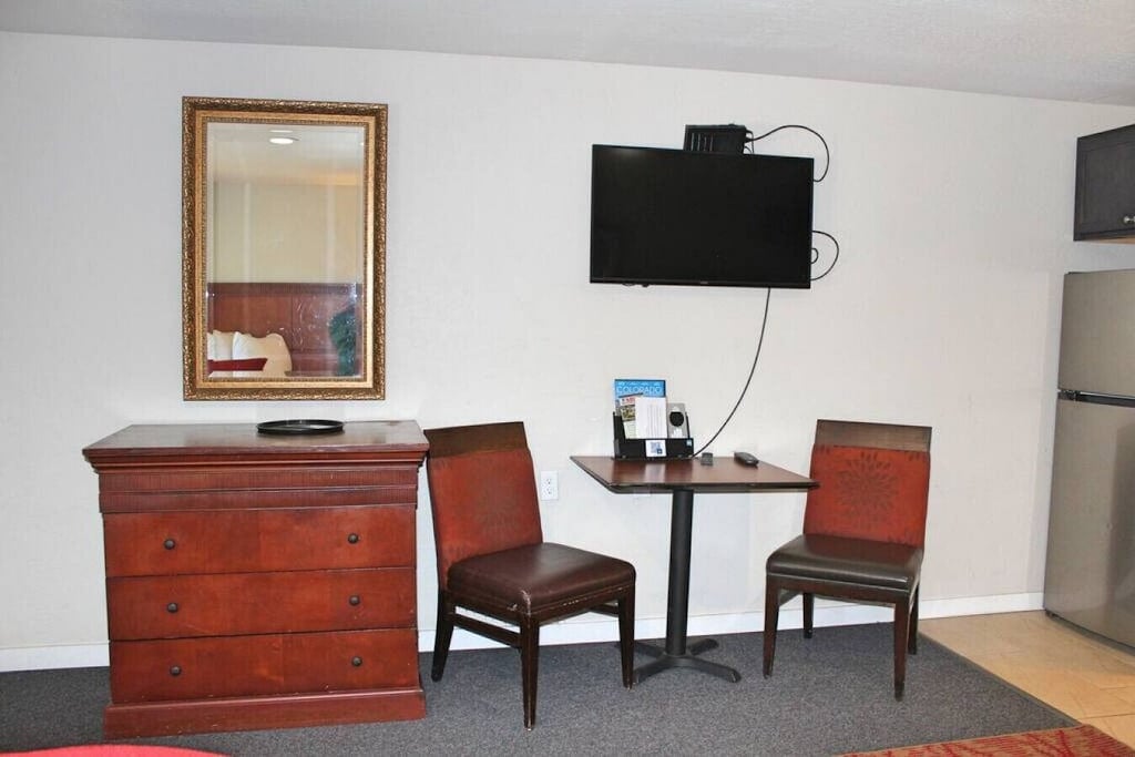 A compact seating area features a wooden table accompanied by two chairs. A chest of drawers is positioned nearby, and a flat-screen television is mounted on the wall above. A mirror adds depth to the space, reflecting soft, natural light.