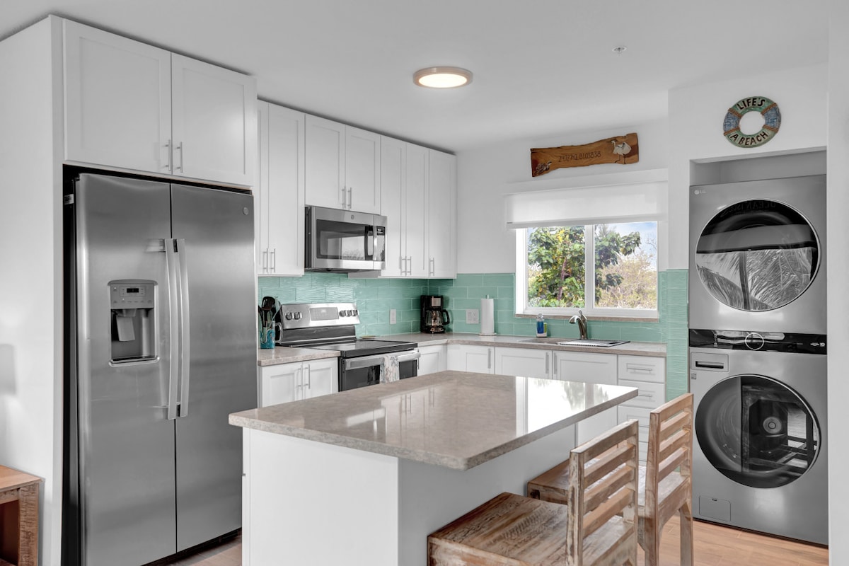 A modern kitchen is equipped with stainless steel appliances, including a refrigerator and microwave. Light wood cabinetry sits above turquoise backsplash tiles. A spacious island serves as a meal prep area, complemented by wooden stools. A window offers natural light and views of greenery outside.