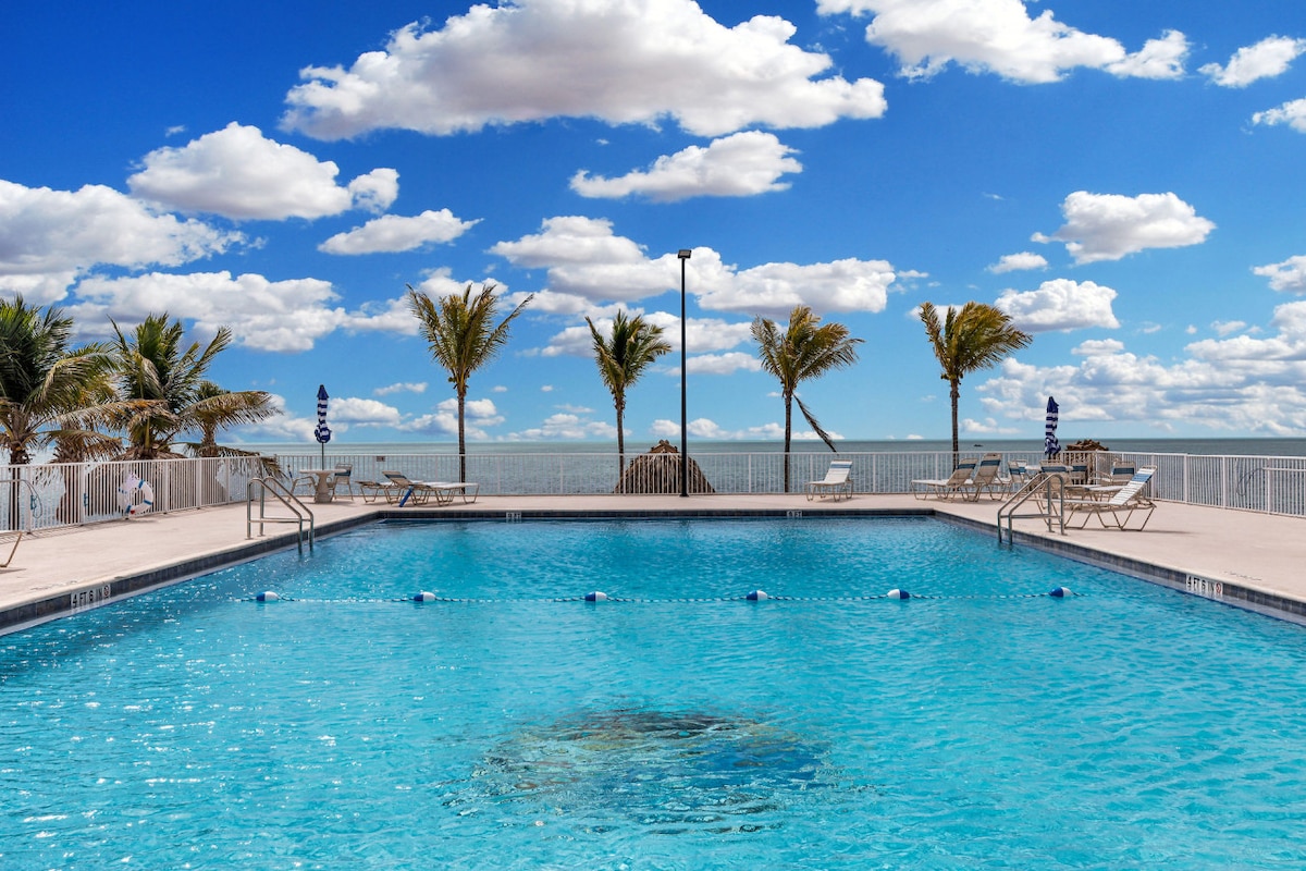 An expansive swimming pool is situated against a backdrop of bright blue sky and fluffy white clouds. Palms sway gently on the pool's perimeter, with several lounge chairs placed for relaxation. The inviting water reflects the colors of the sky, enhancing the tranquil atmosphere.