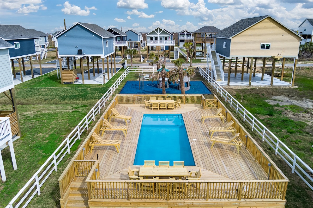 An outdoor community pool area is surrounded by wooden decking, with multiple lounge chairs positioned for relaxation. The pool is centrally located among stilted villas, offering a playful playground nearby, set within an open grassy space against a backdrop of blue skies.