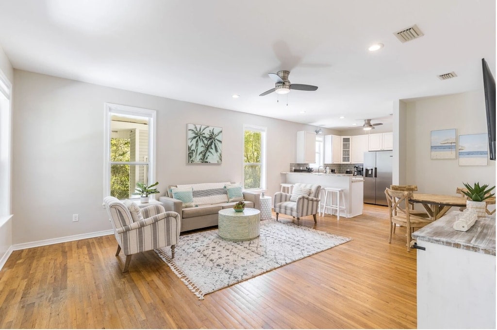 A spacious living area features light wood flooring and ample natural light. Comfortable seating options include a plush sofa and accent chairs. A decorative area rug lies beneath a central coffee table, with views leading to the dining area and updated kitchen in the background.