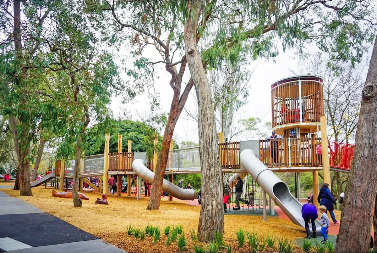 A vibrant playground is surrounded by natural trees, featuring wooden structures and multiple slides. Children can be seen playing on the equipment, while a sandy area is present underneath. The setting offers a spacious environment for outdoor activities and social interaction.