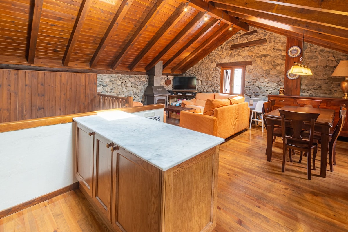 An open-plan kitchen and living area features wooden beams and a stone wall, creating a cozy ambiance. A marble countertop divides the kitchen from the space, where a dining table and chairs accommodate four people. A sofa and TV are positioned for relaxation.