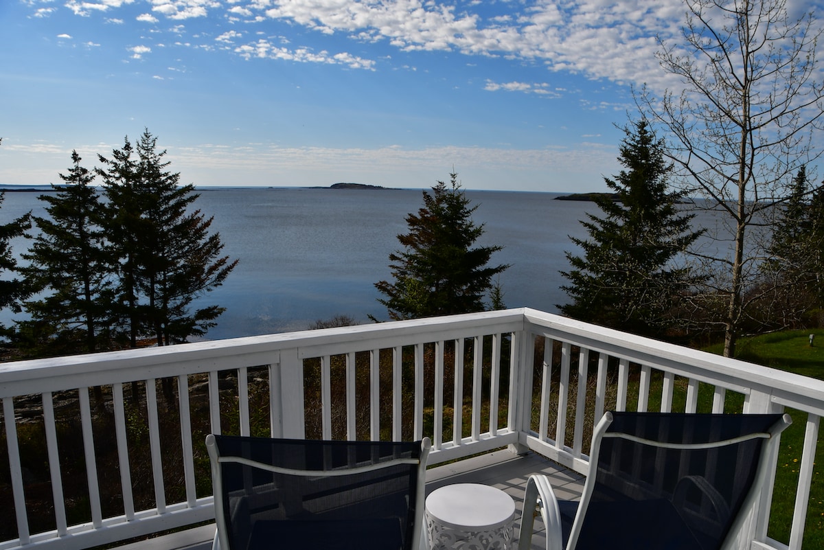 A serene view of the coastline is displayed from a deck featuring two chairs and a small table. The calm water reflects the sky, while trees line the shore, creating a peaceful setting perfect for relaxation.