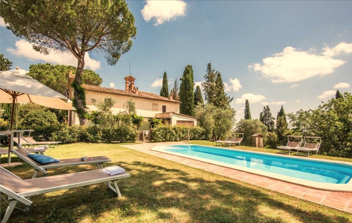 A swimming pool is framed by a lush garden, featuring lounge chairs placed on the grass. A large tree offers shade nearby. The villa, partially visible in the background, is surrounded by tall greenery, with a clear sky and soft clouds above.