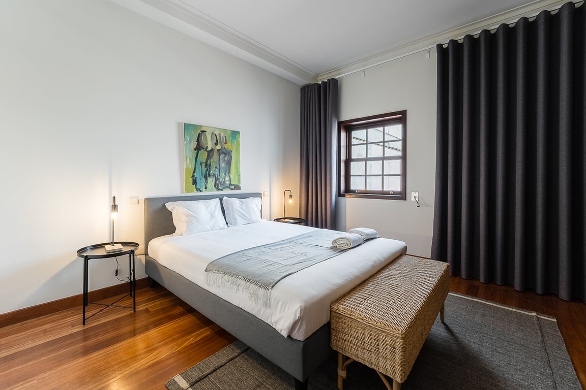 A bright bedroom features a spacious bed, crisp white linens, and a minimalist design. A large window offers natural light, partially shaded by dark curtains. A modern artwork adds a touch of color, while a wicker bench provides additional seating.