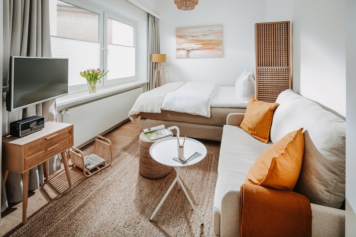 The combined living and sleeping area features a spacious bed with crisp white linens. A neutral-toned sofa with accent pillows sits adjacent to a small coffee table. Natural light enters through large windows, enhancing the warm ambiance of the room.