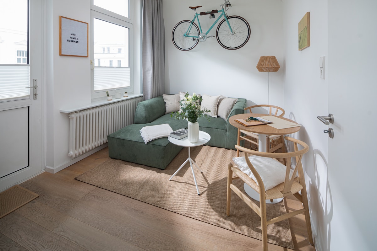 A welcoming living area is presented with a soft green sectional sofa and a circular white coffee table. Natural light floods in through a large window, while a bicycle decoratively hangs on the wall. Two wooden chairs, accompanied by a small desk, create a cozy nook.