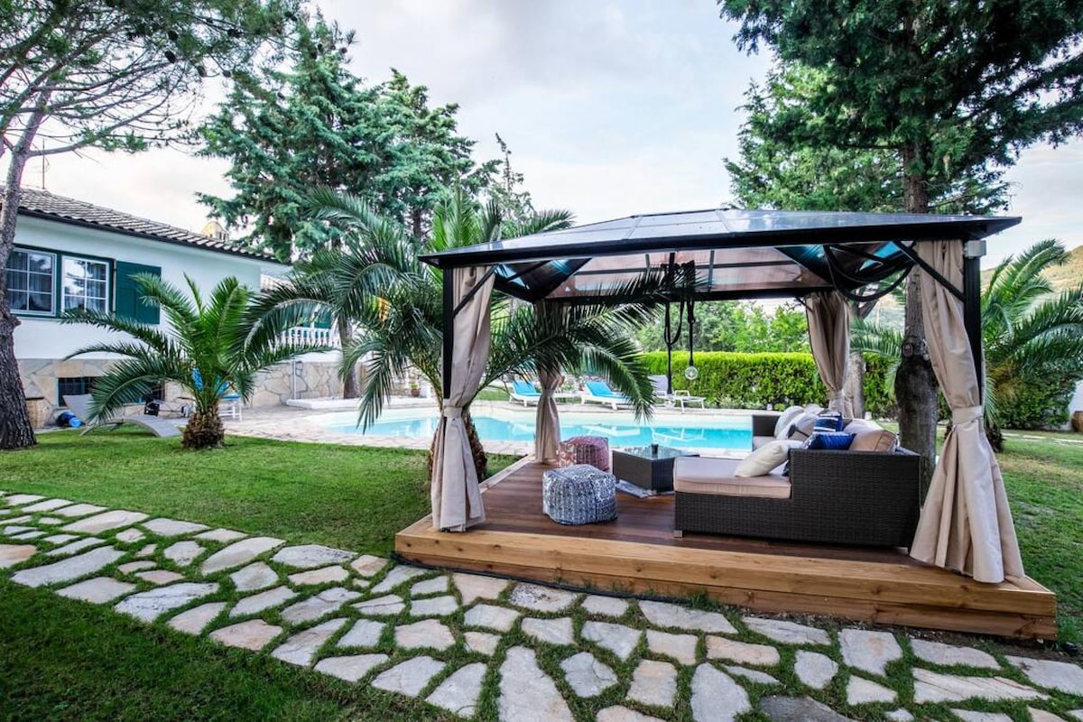 A shaded gazebo with linen drapes offers a comfortable seating area, situated on a wooden deck. Lush greenery surrounds the space, and a clear swimming pool is visible in the background, providing a tranquil atmosphere for relaxation.