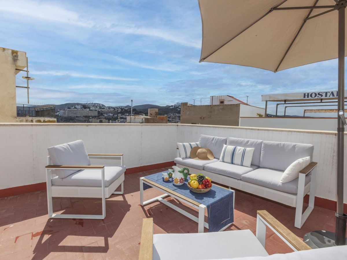 A spacious terrace is furnished with a white couch and armchairs, offering comfortable seating. A small table is set with fresh fruit and a blue cloth, while a large umbrella provides shade. Scenic views of the surrounding area are visible in the background.