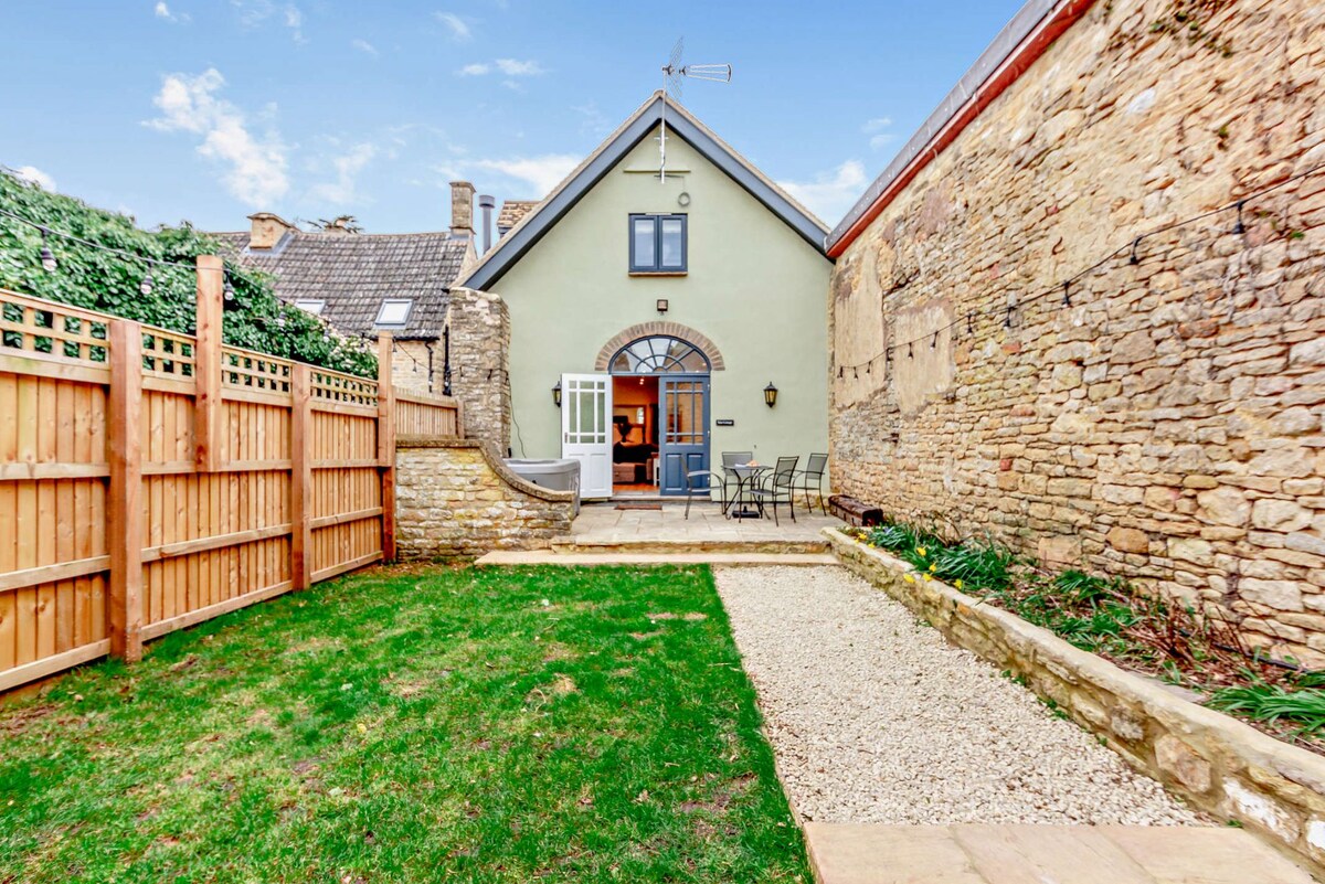 The private garden features a gravel path leading to a stone patio area, framed by a lush green lawn. The cottage's back entrance is visible, along with outdoor seating for four and a wooden fence providing privacy. Stone walls and greenery create a tranquil backdrop.