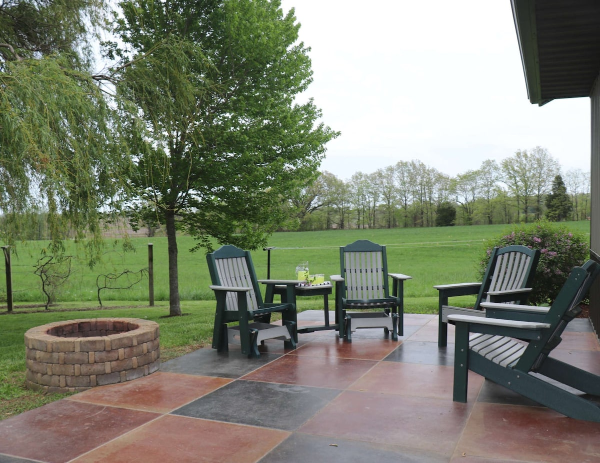 A patio area features four outdoor chairs arranged around a small table, with a firepit made of stone nearby. Lush greenery surrounds the space, offering a view of the expansive countryside. A large tree provides shade, enhancing the outdoor setting.