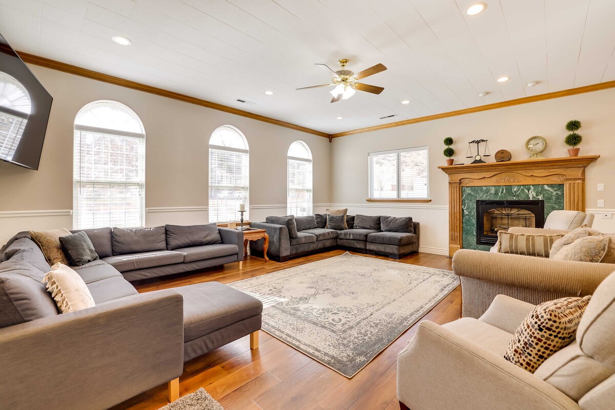 A spacious living room features multiple sectional sofas arranged around a large area rug. Natural light filters through arched windows, illuminating the hardwood floor. A decorative fireplace with a green marble surround creates an inviting focal point, complemented by a ceiling fan overhead.