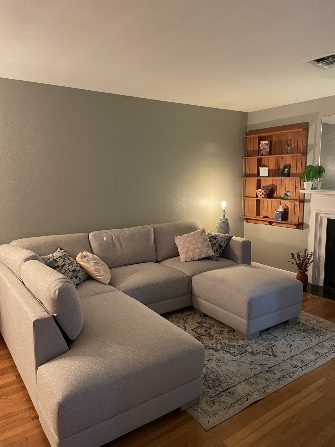 A light gray sectional sofa is positioned in a corner, complemented by decorative pillows. A small ottoman is nearby. A wooden shelf with various decorative items is mounted on the wall, and a subtle light source creates a warm ambiance within the space.